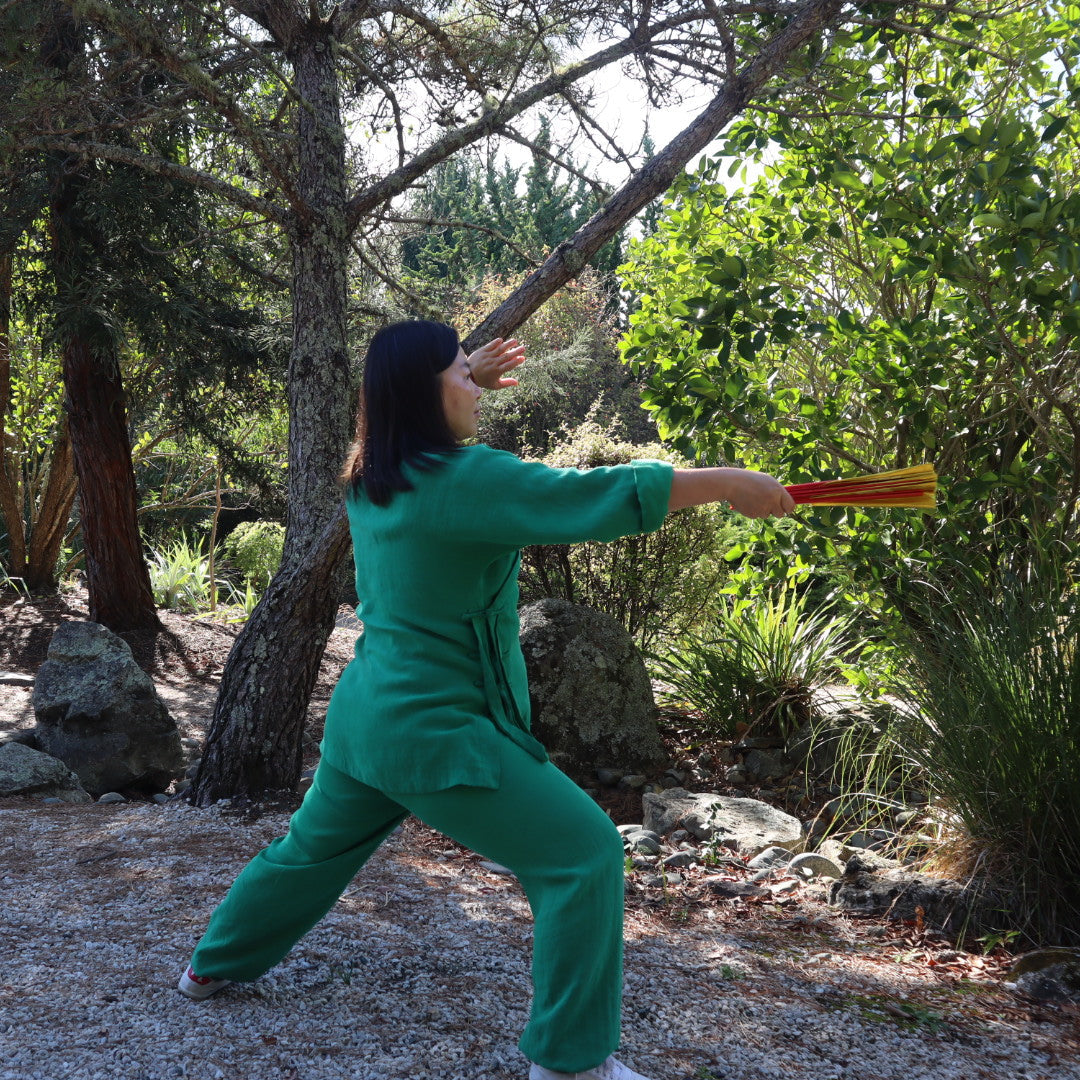 A woman in a green martial arts uniform practices a sword form outdoors in a wooded area with trees, rocks, and greenery.