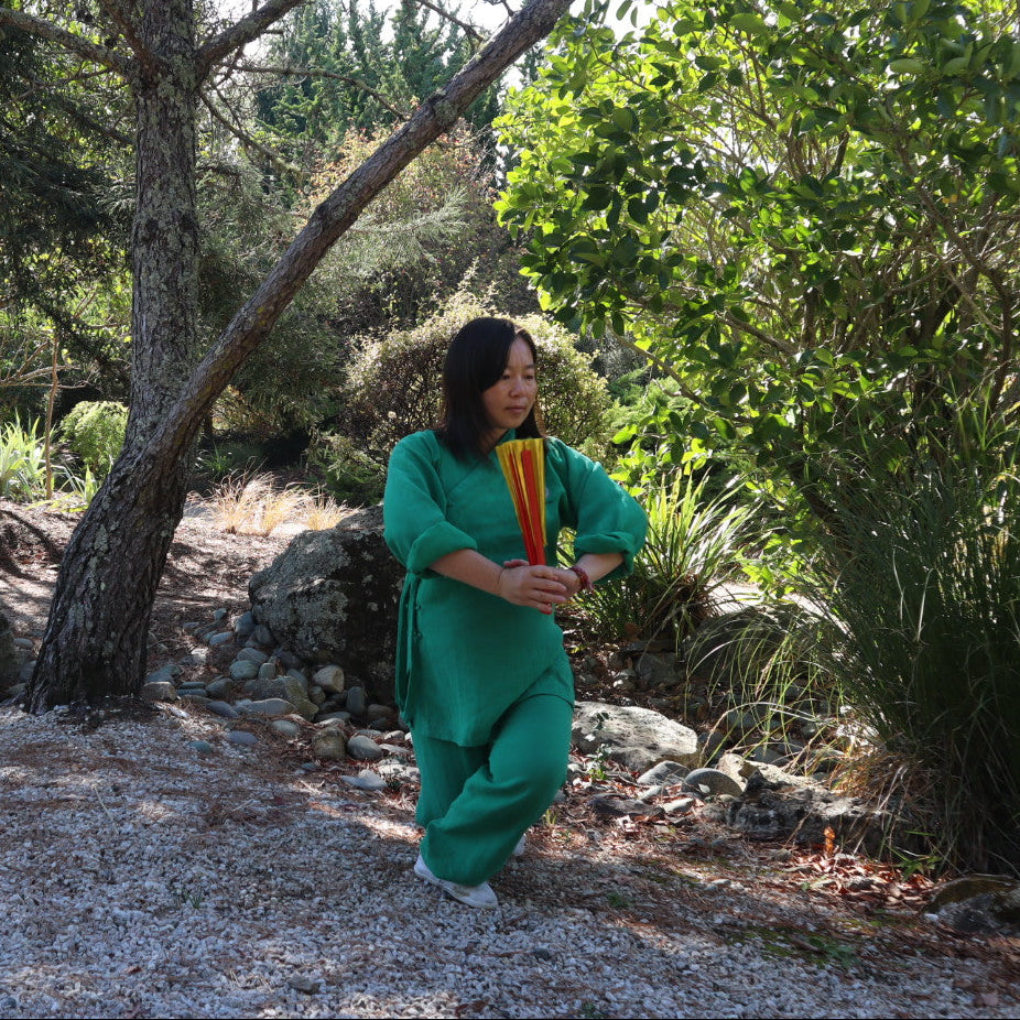 A woman wearing a bright green full-body outfit stands outdoors in a wooded area with trees, rocks, and gravel ground, holding a yellow and orange striped item in front of her.