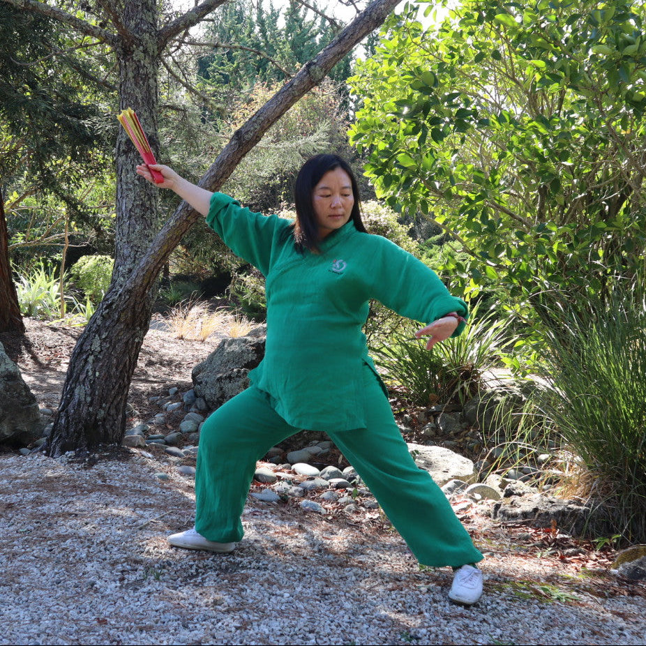 A woman in a green martial arts uniform practices a stance outdoors in a wooded area, holding a wooden practice sword.
