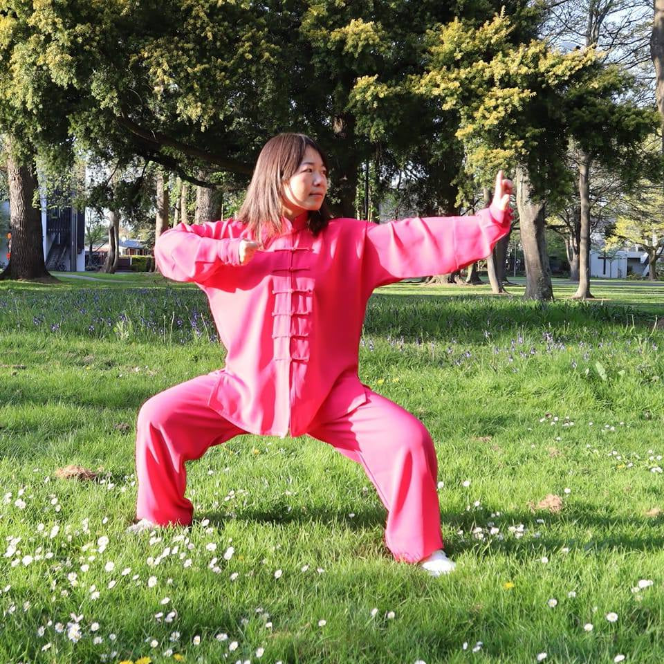 Person in a pink outfit practicing tai chi in a park with trees and grass.